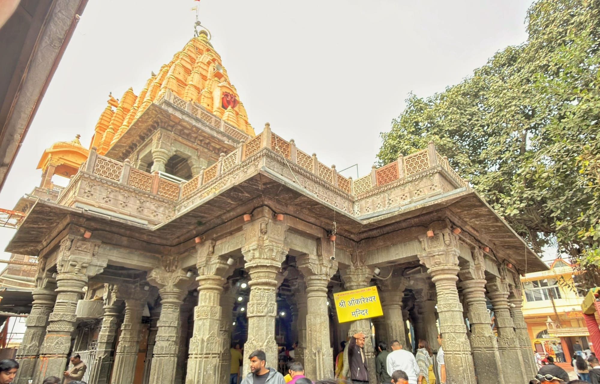 The orange, tiered spire of the Mahakaleshwar temple rising above a pillared hall of carved grey stone, with people standing between the pillars