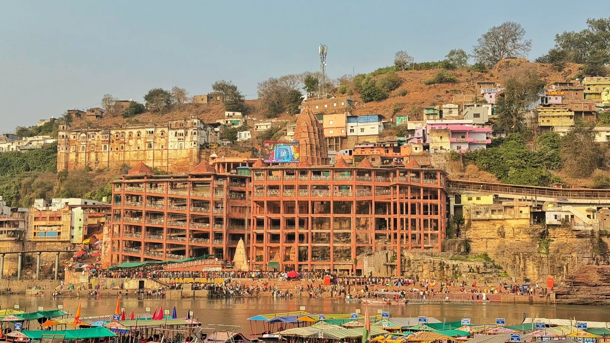 The ghats of Omkareshwar seen across the river, with terraces of orange buildings and temples climbing a hillside and boats and a crowd at the water's edge